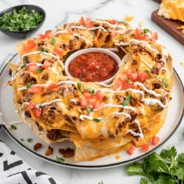 A ring of cheesy Bundt Pan Nachos with ground beef, sour cream, chopped tomatoes, and cilantro surrounds a bowl of salsa on a white plate. Shredded cheese, cilantro, and diced tomatoes are nearby on a marble surface.