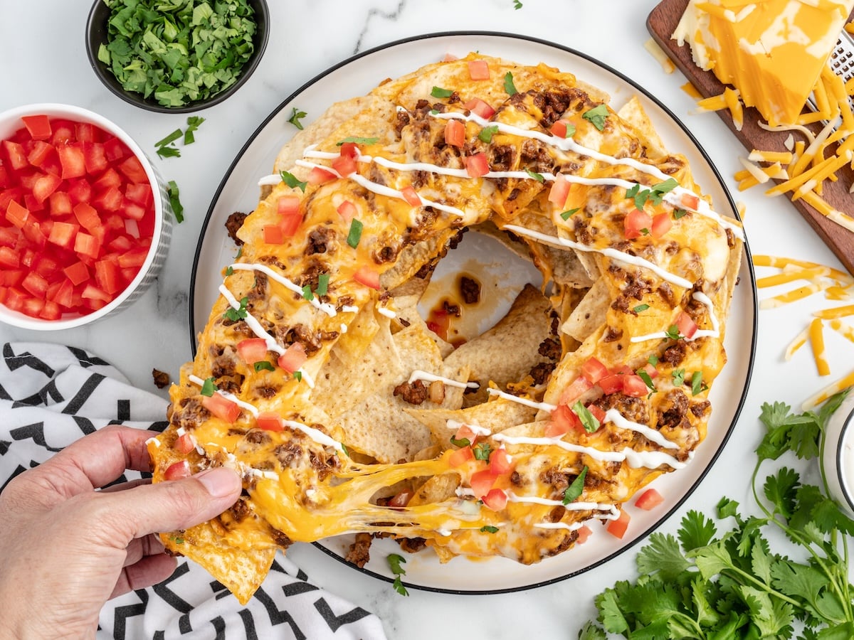 A round plate of Bundt Pan Nachos arranged in a ring shape, topped with melted cheese, ground beef, diced tomatoes, and drizzled with sour cream. A hand pulls out a cheesy nacho. Bowls of tomatoes and cilantro sit nearby.
