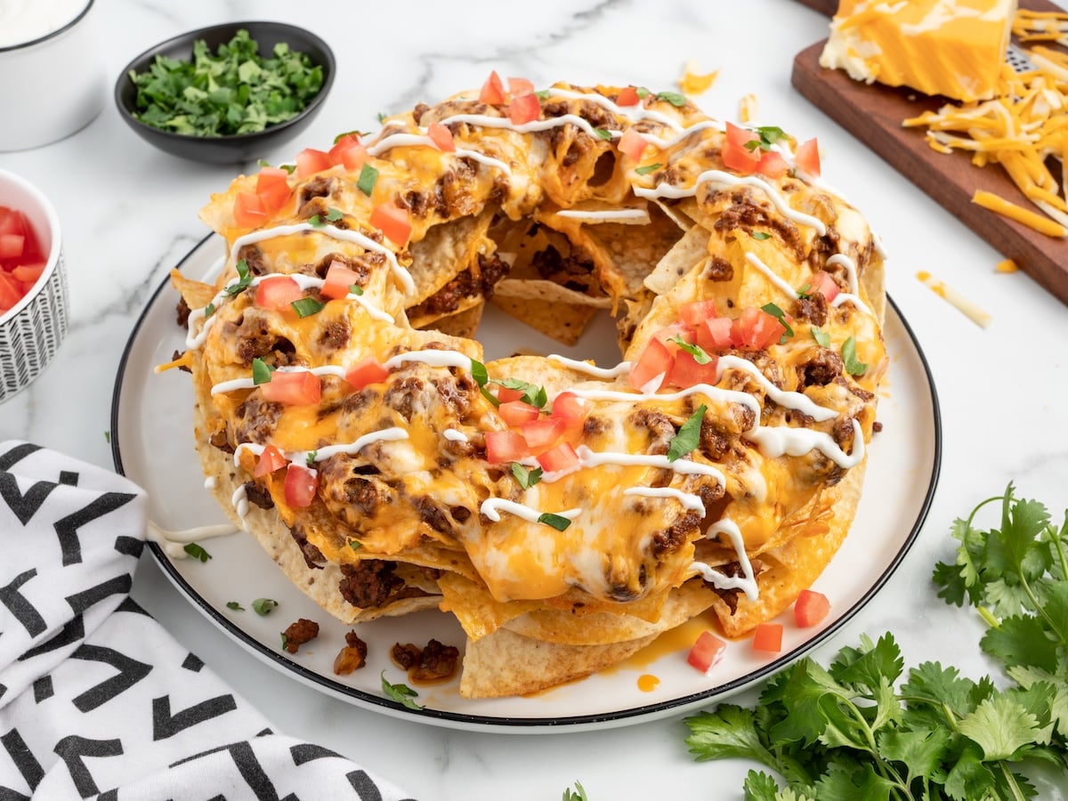 A round platter of Bundt Pan Nachos arranged in a ring, topped with melted cheese, seasoned ground beef, diced tomatoes, chopped green herbs, and drizzled with sour cream. Bowls of garnishes and ingredients are nearby.