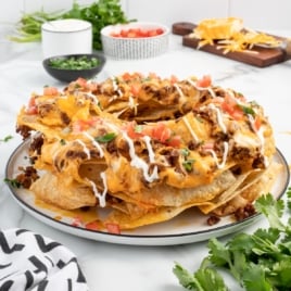 A ring-shaped stack of Bundt Pan Nachos topped with melted cheese, seasoned ground beef, diced tomatoes, chopped cilantro, and a drizzle of sour cream, served on a white plate with fresh ingredients in the background.