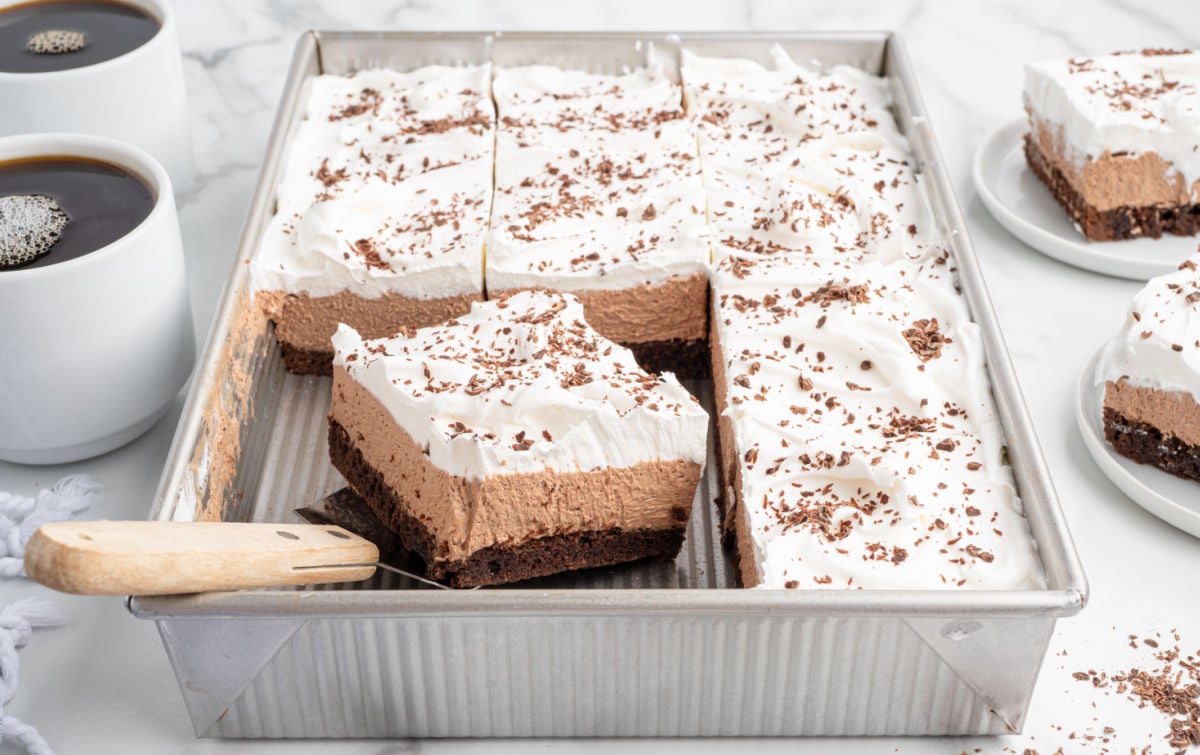 A metal baking pan filled with sliced French silk brownies topped with whipped cream and chocolate shavings. A serving spatula lifts one square, showing the distinct layers of brownie and mousse. Coffee cups sit beside the pan.