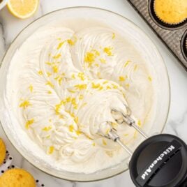 A mixer blends Lemon Cream Cheese Frosting with lemon zest in a glass bowl, surrounded by fresh lemons, yellow cupcakes, and a muffin tin on a marble surface.