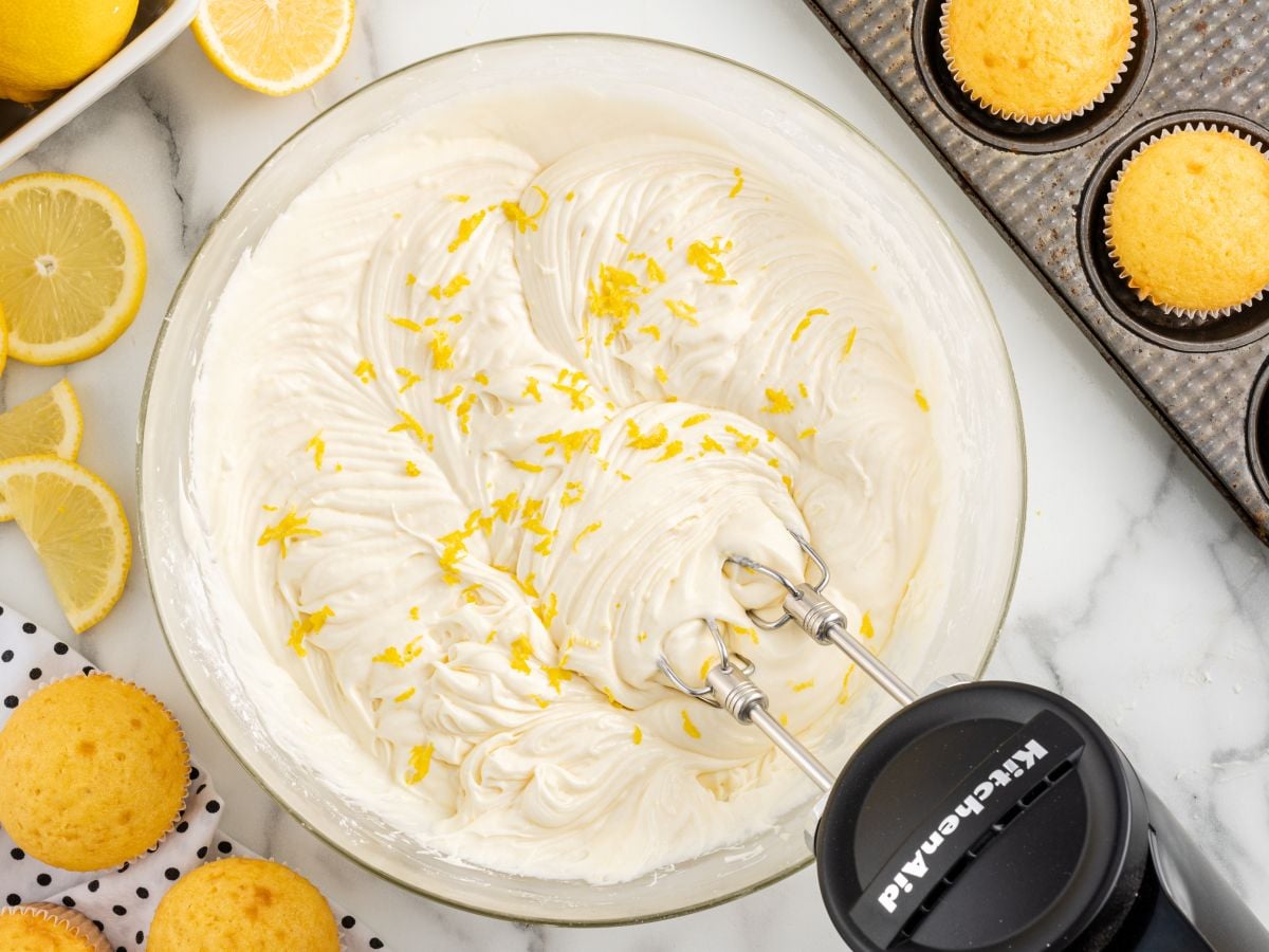 A mixer blends Lemon Cream Cheese Frosting with lemon zest in a glass bowl, surrounded by fresh lemons, yellow cupcakes, and a muffin tin on a marble surface.