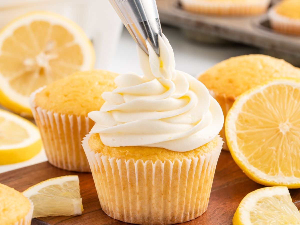 A close-up of a lemon cupcake being topped with swirled Lemon Cream Cheese Frosting from a piping bag, surrounded by lemon slices and more cupcakes on a wooden board.
