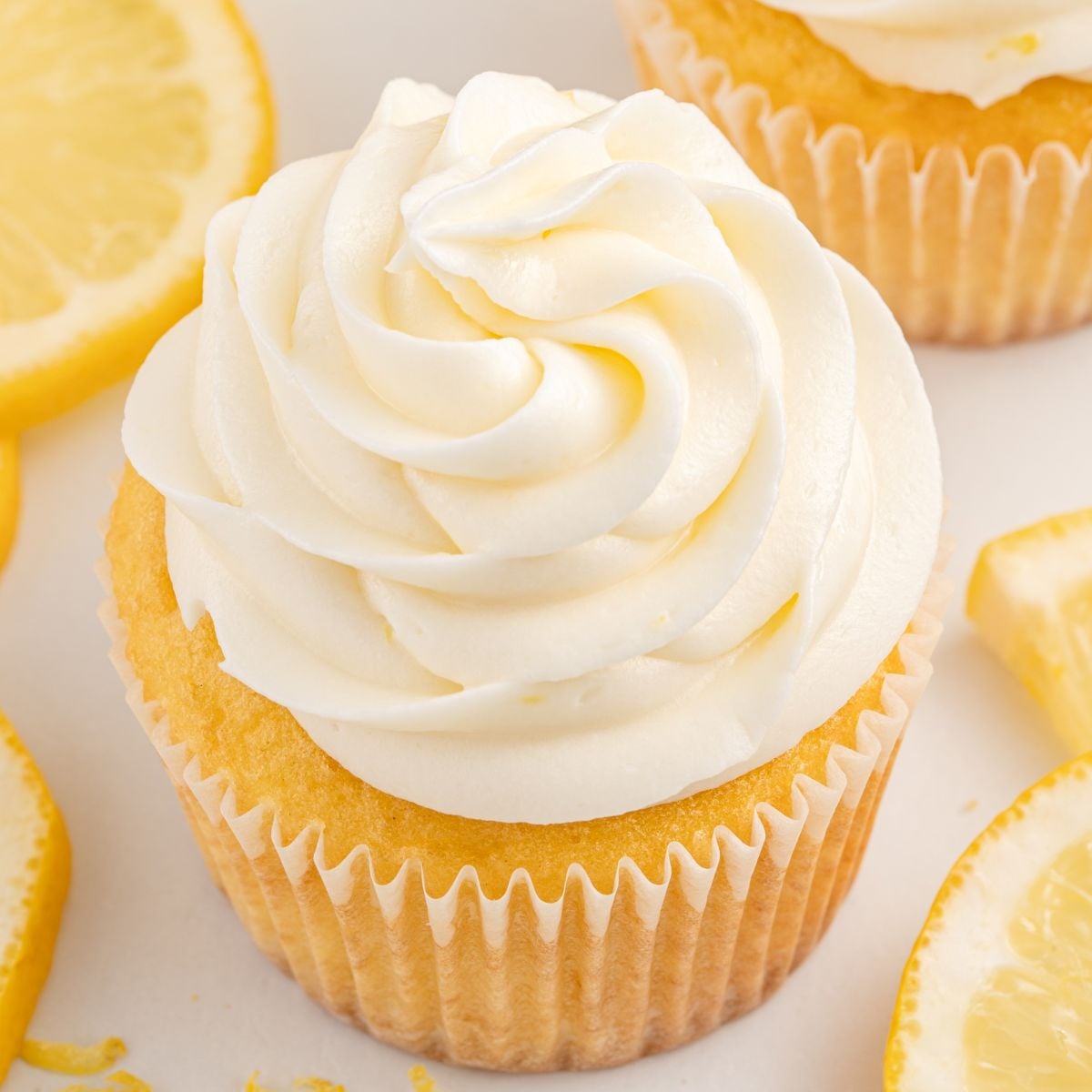 A lemon cupcake with a swirl of luscious Lemon Cream Cheese Frosting on top, surrounded by several slices of fresh lemon.