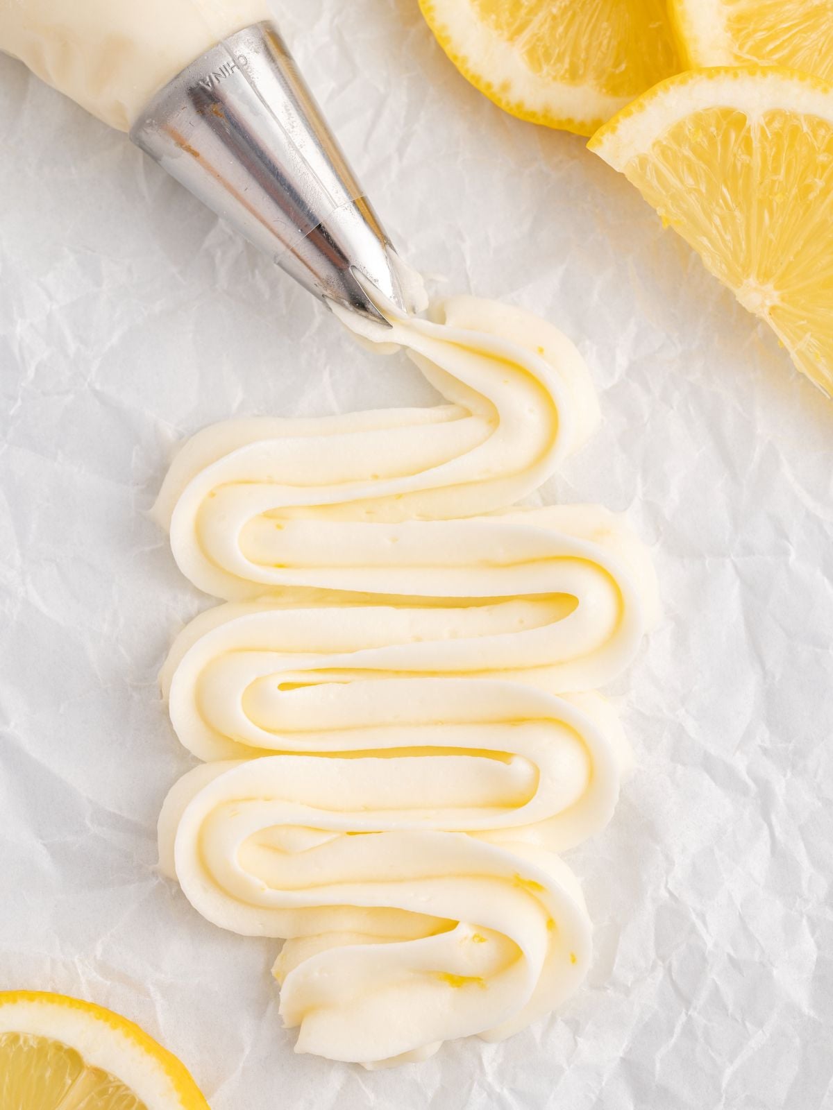 A metal piping tip spreads a zigzag line of Lemon Cream Cheese Frosting with lemon zest on crumpled white parchment paper, surrounded by fresh lemon slices.