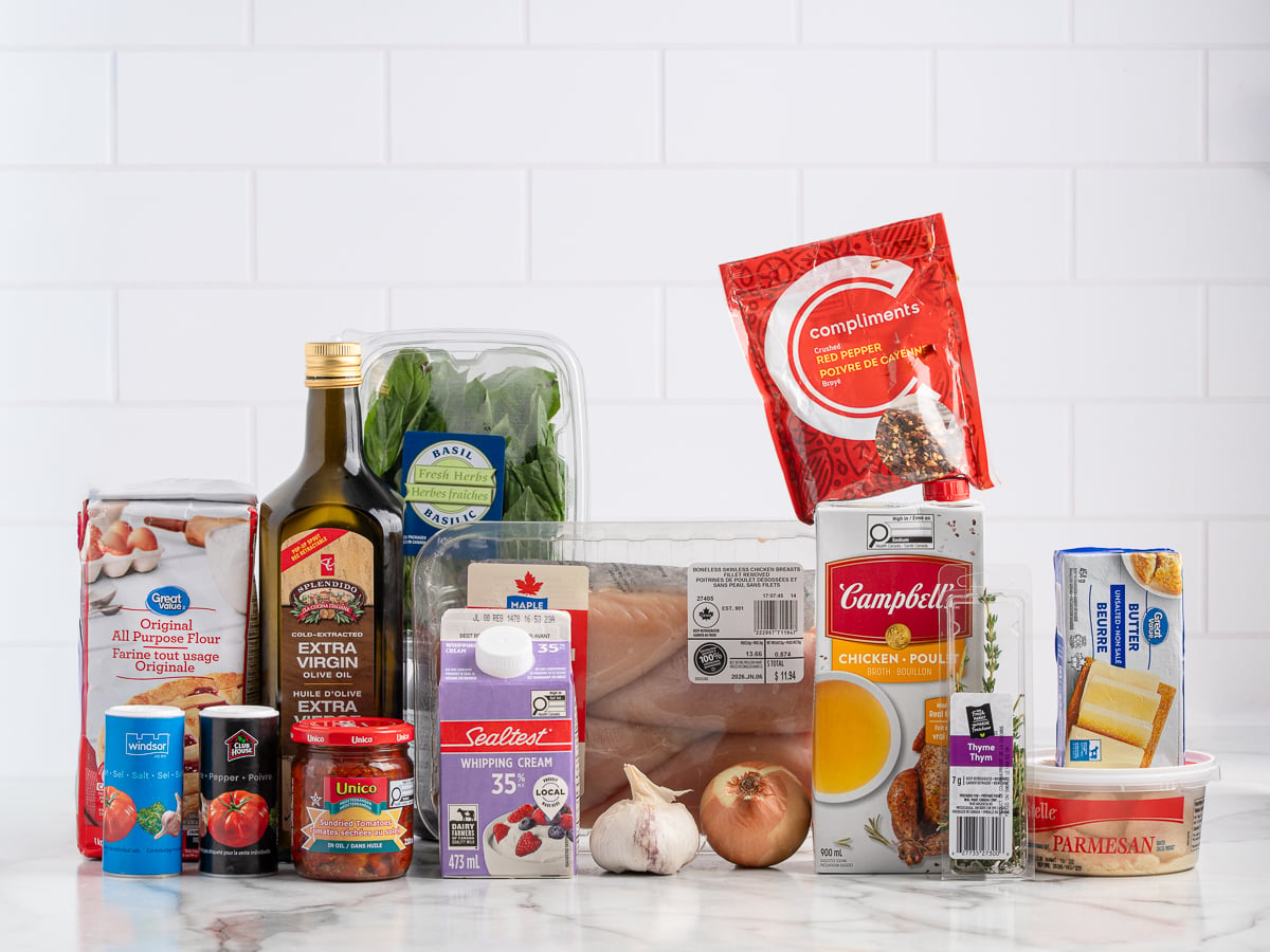 Assorted groceries on a counter for Marry Me Chicken, including chicken breasts, olive oil, canned tomatoes, flour, parmesan cheese, whipping cream, chicken broth, spinach, butter, onion, garlic, and seasonings, set against a white tile background.