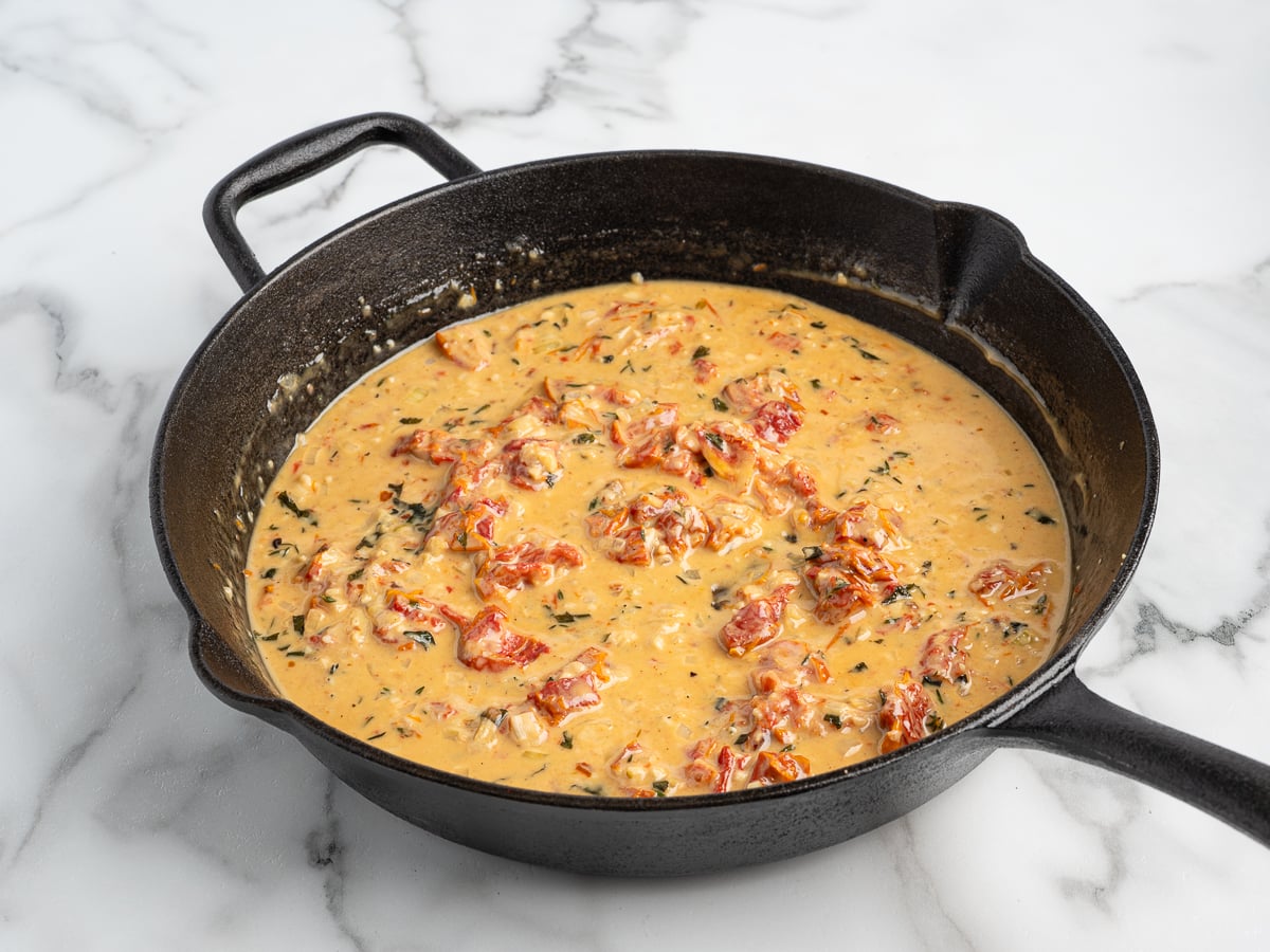 A black cast iron skillet on a marble surface filled with creamy, orange-colored Marry Me Chicken sauce featuring chopped sun-dried tomatoes and fresh herbs.