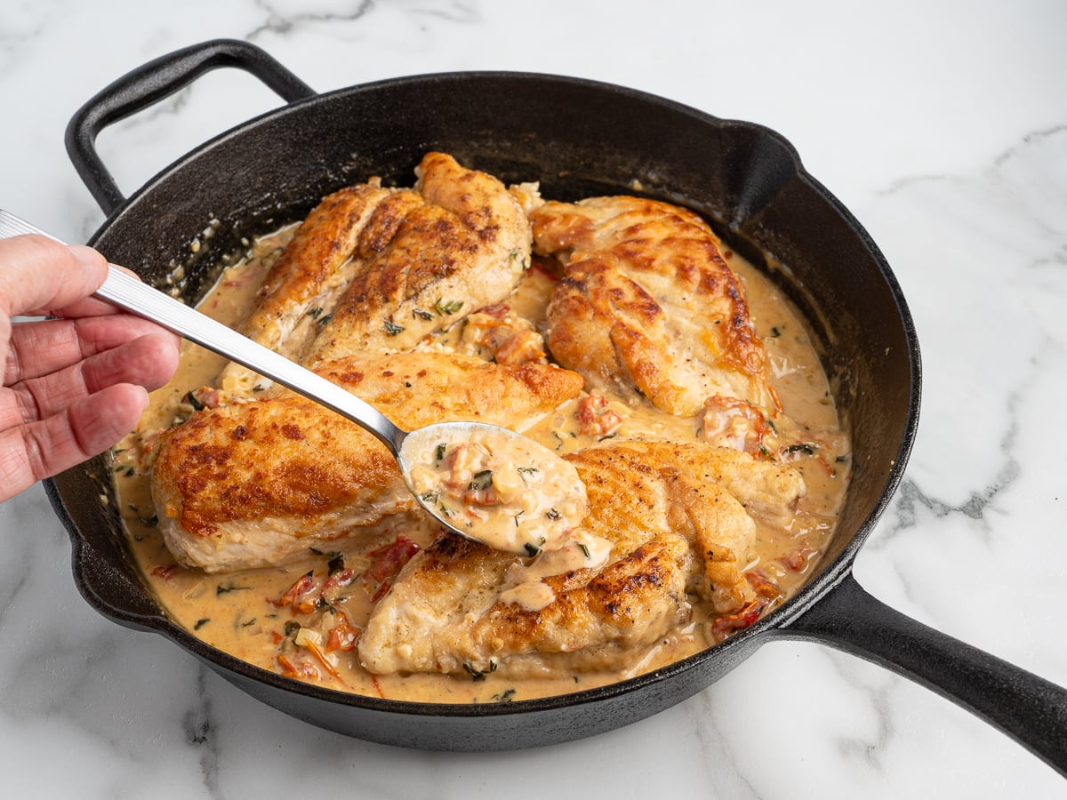 A hand holds a spoon, serving creamy Marry Me Chicken sauce over browned chicken breasts in a black cast-iron skillet. The sauce features herbs and chopped vegetables. The skillet rests on a white marble surface.