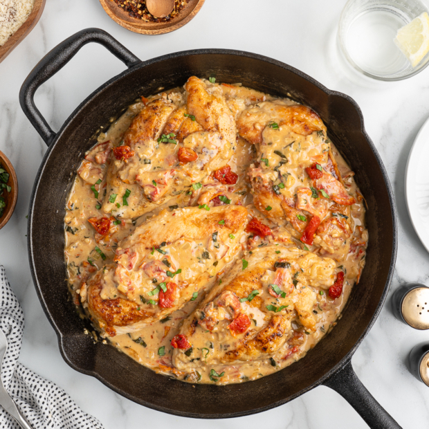 A cast iron skillet filled with creamy Marry Me Chicken breasts in a rich sauce with spinach and sun-dried tomatoes, surrounded by fresh herbs, utensils, and condiments on a white marble surface.