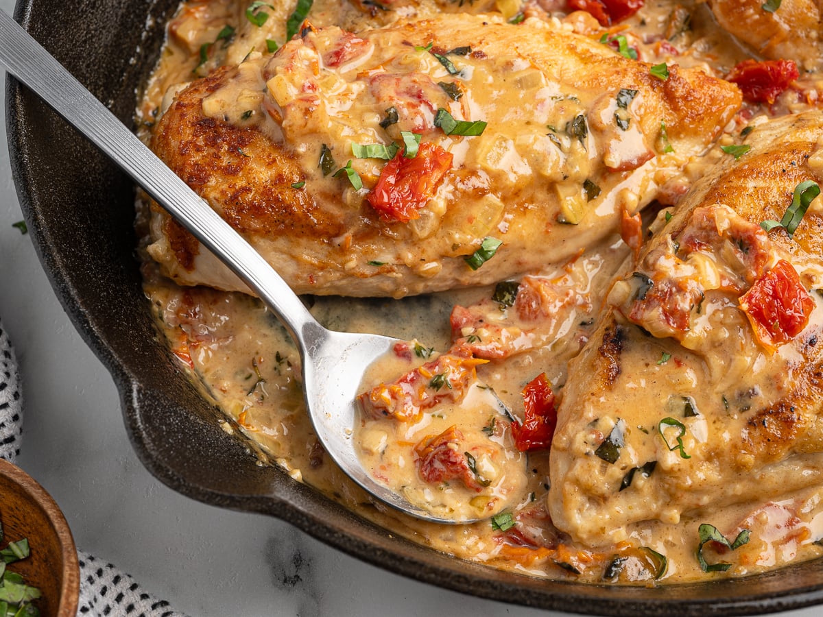 A close-up of creamy Marry Me Chicken breasts in a skillet, topped with rich garlic sauce, sun-dried tomatoes, and fresh herbs, with a spoon resting beside the chicken.