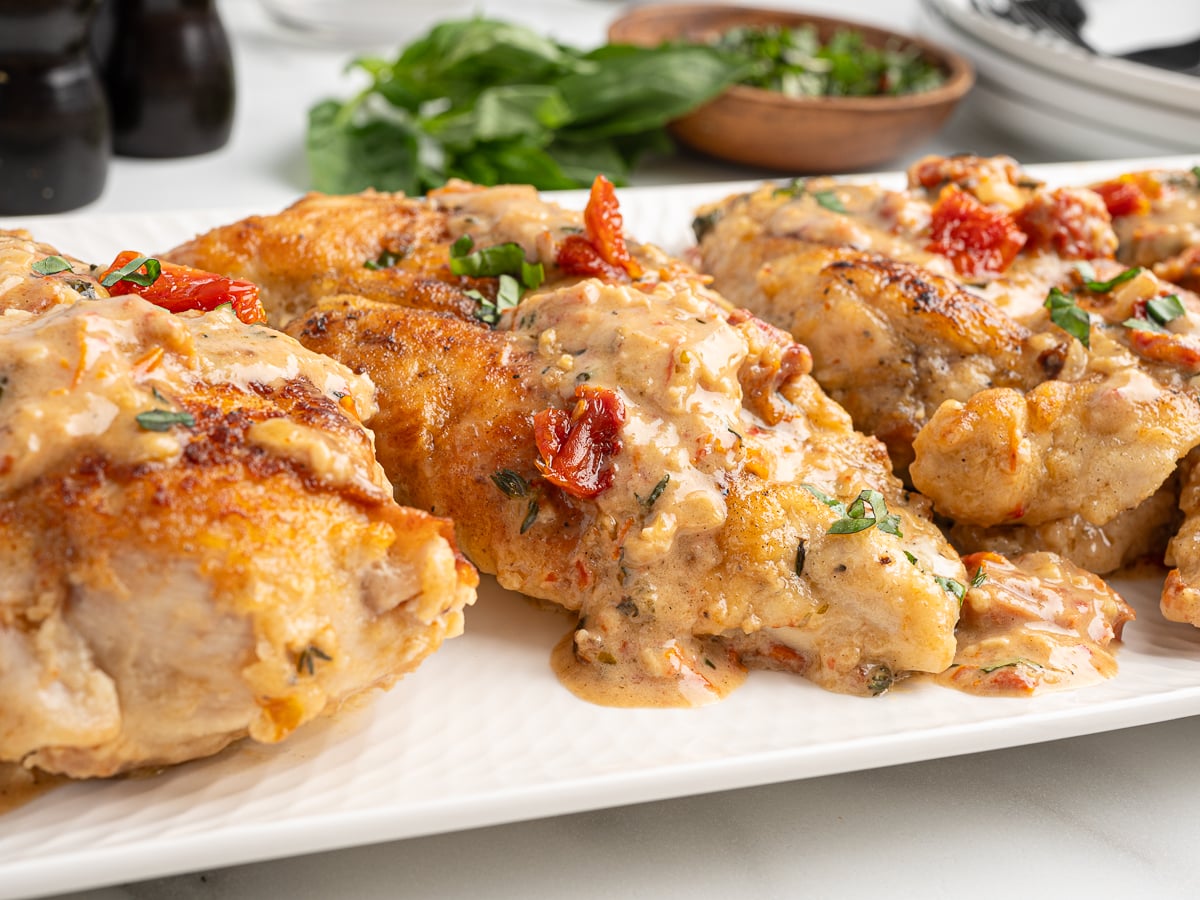 Three pieces of creamy Marry Me Chicken breast, garnished with chopped herbs and sun-dried tomatoes, are served on a white platter, with fresh greens and bowls of herbs in the background.