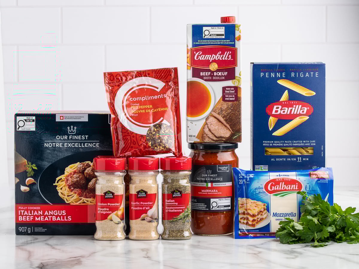 Various packaged food items arranged on a marble surface, including beef meatballs, dried herbs, chili powder, beef broth, marinara sauce, penne pasta, shredded mozzarella, and fresh parsley—perfect for a hearty Meatball Casserole.