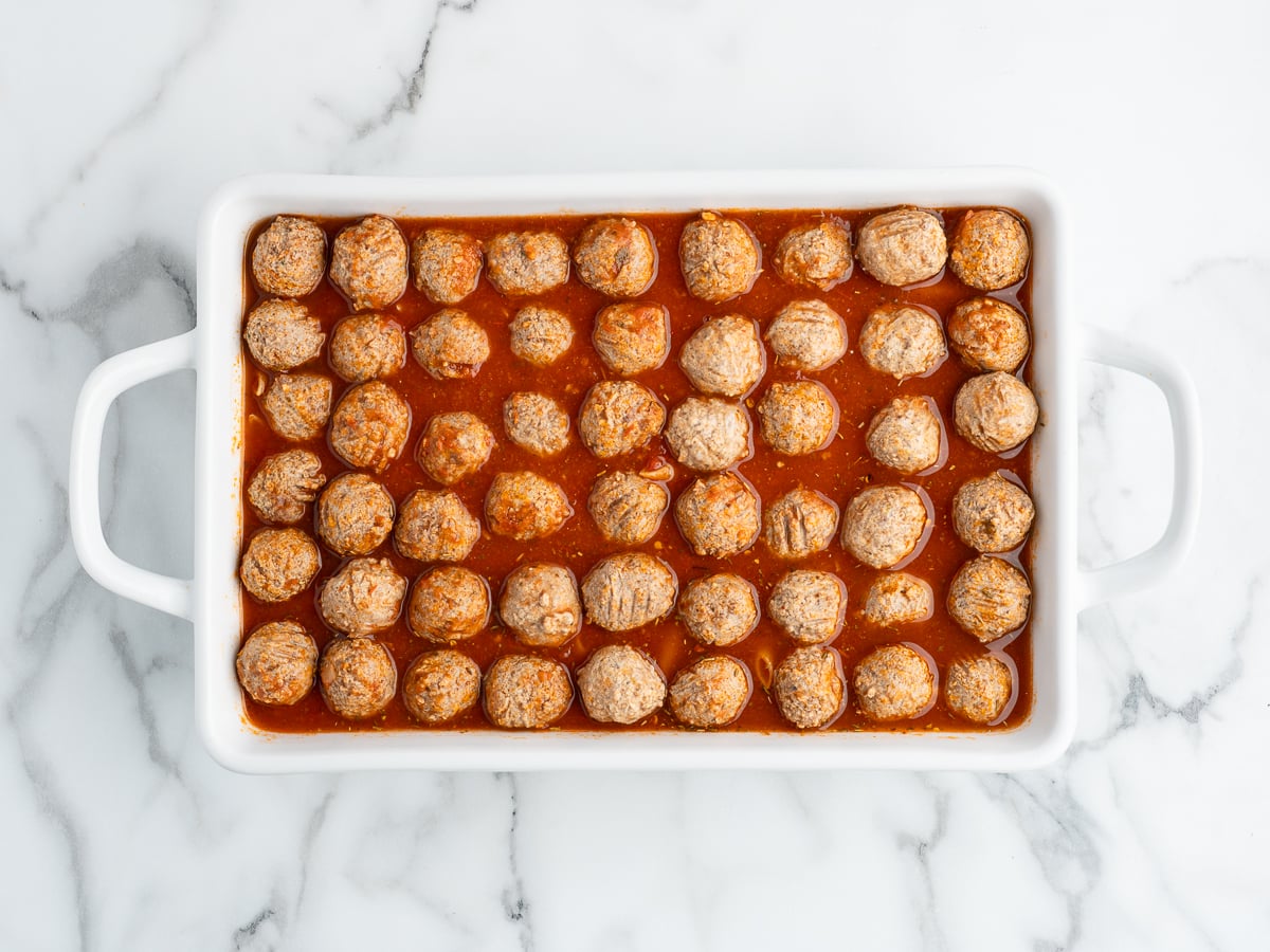 A white rectangular baking dish filled with rows of savory Meatball Casserole in red tomato sauce, placed on a white marble surface.