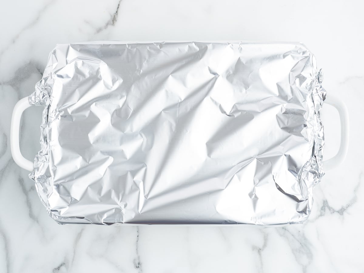 A rectangular white baking dish, likely holding a savory Meatball Casserole, is covered with crinkled aluminum foil and sits on a white marble surface.