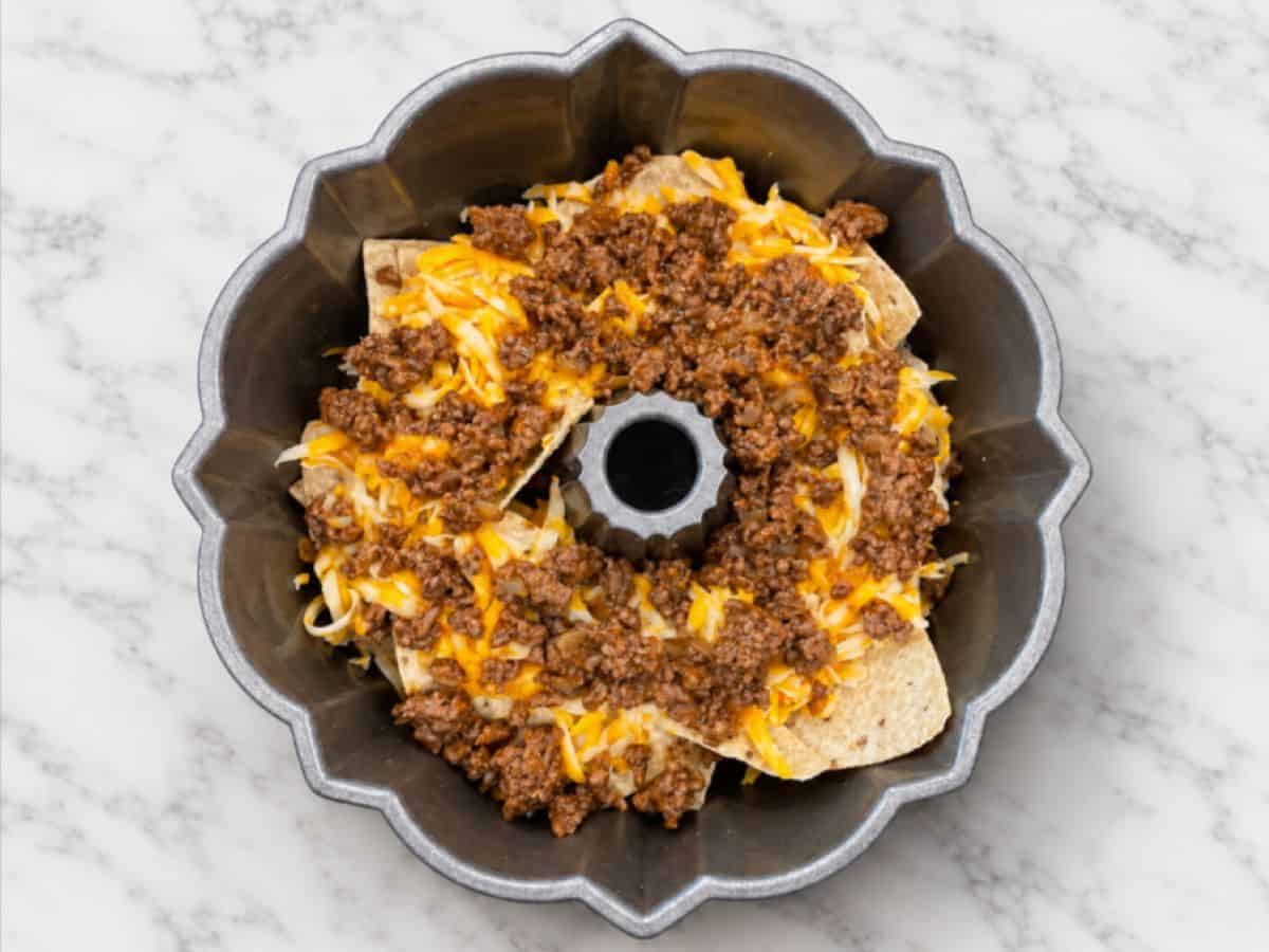 A Bundt Pan Nachos masterpiece sits on a marble surface, filled with irresistible layers of tortilla chips, seasoned cooked ground beef, and gooey melted shredded cheese.