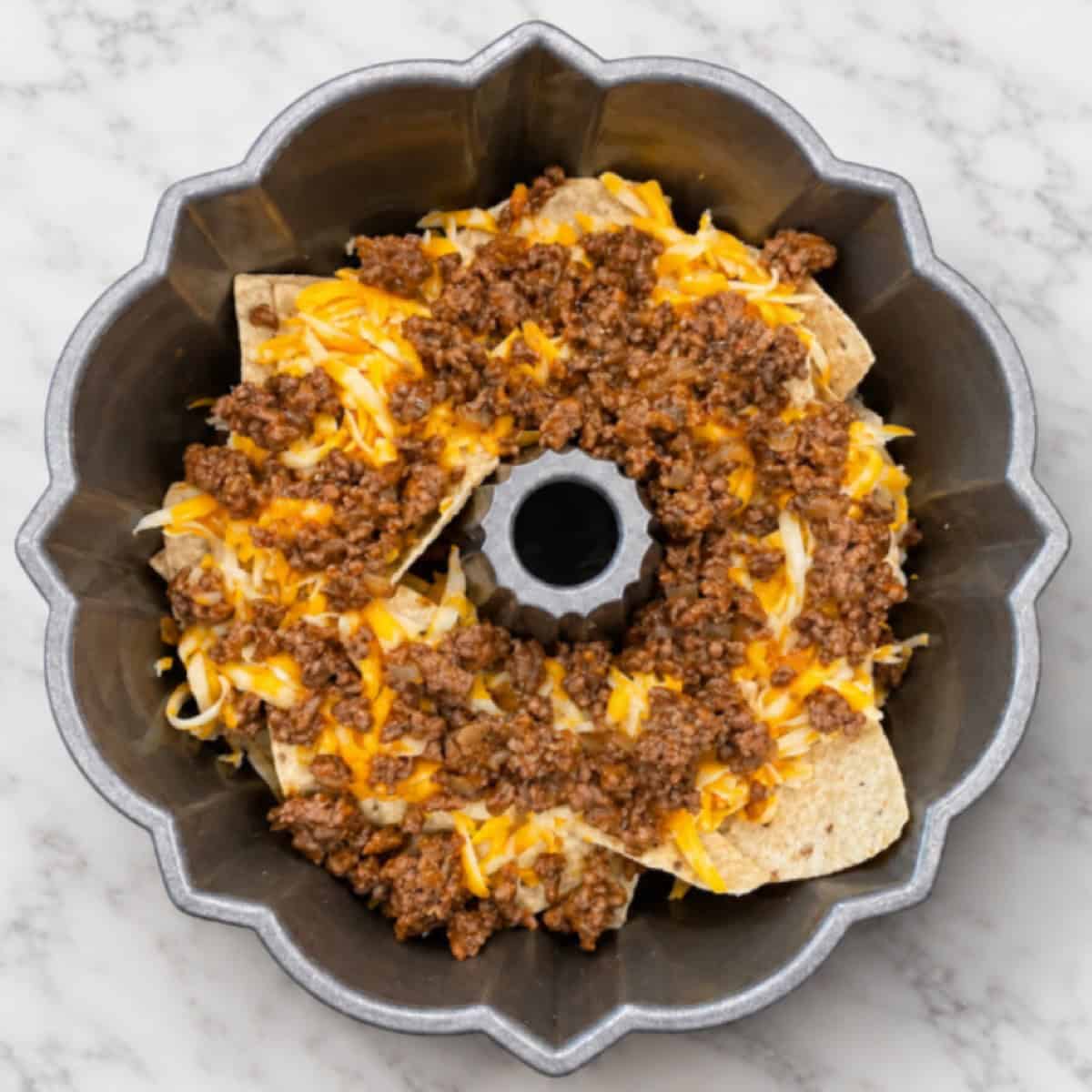 Bundt Pan Nachos come to life in this creation—a Bundt pan filled with layers of tortilla chips, seasoned ground beef, and shredded cheese, all arranged in a circular pattern on a marble countertop.
