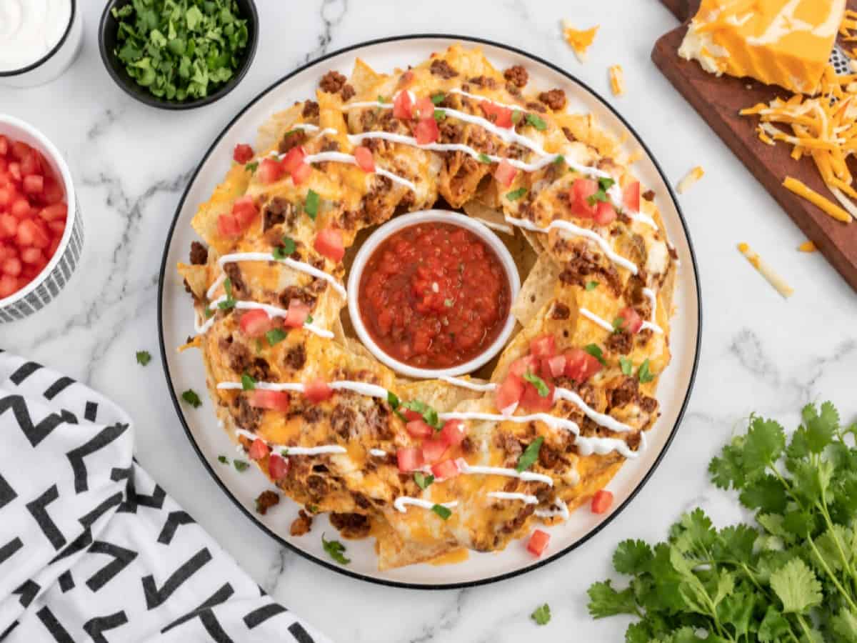 A plate of Bundt Pan Nachos arranged in a ring around a bowl of salsa, topped with melted cheese, diced tomatoes, ground meat, and drizzled with sour cream, with cilantro and chopped green onions nearby.