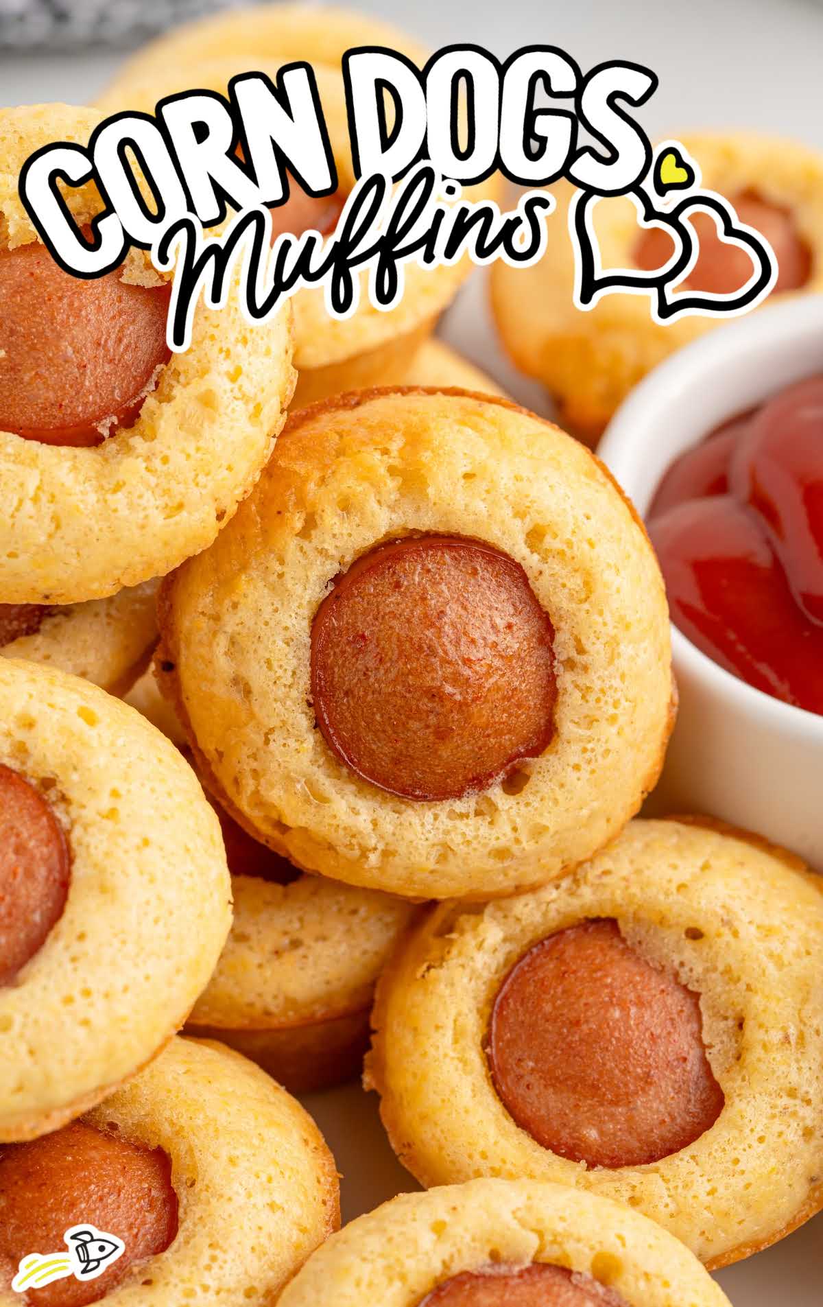 Corn dog muffin dipped in ketchup with other muffins blurred in the background.