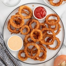 Overhead view of onion rings on a serving plate with dipping sauces, napkin, and utensils