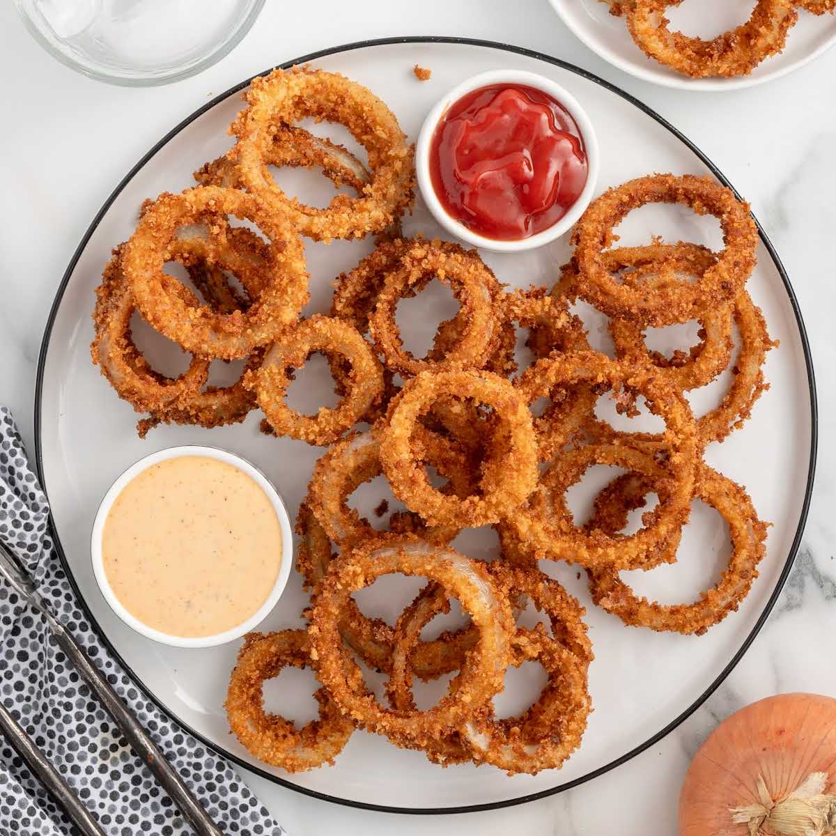 Overhead view of onion rings on a serving plate with dipping sauces, napkin, and utensils
