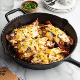 A cast iron skillet filled with Alice Springs Chicken features cooked chicken breasts topped with crispy bacon, melted cheese, and chopped herbs, sitting on a marble countertop beside a small bowl of chopped green herbs.