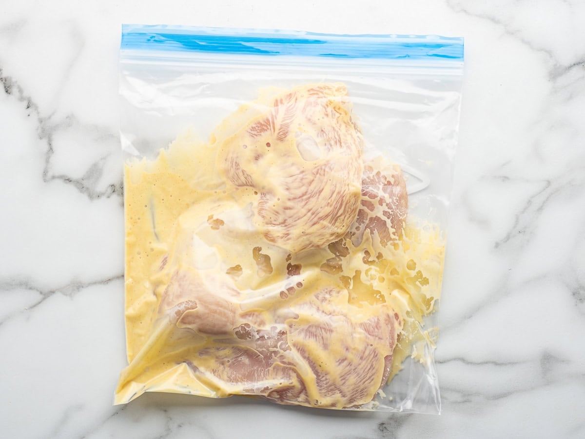 Raw chicken pieces in a resealable plastic bag with yellow marinade, placed on a white marble surface—perfect for preparing Alice Springs Chicken at home.