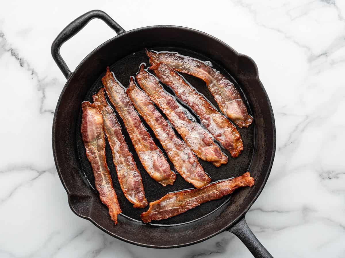 Six strips of crispy bacon, perfect for topping Alice Springs Chicken, cooking in a black cast iron skillet on a white marble surface.