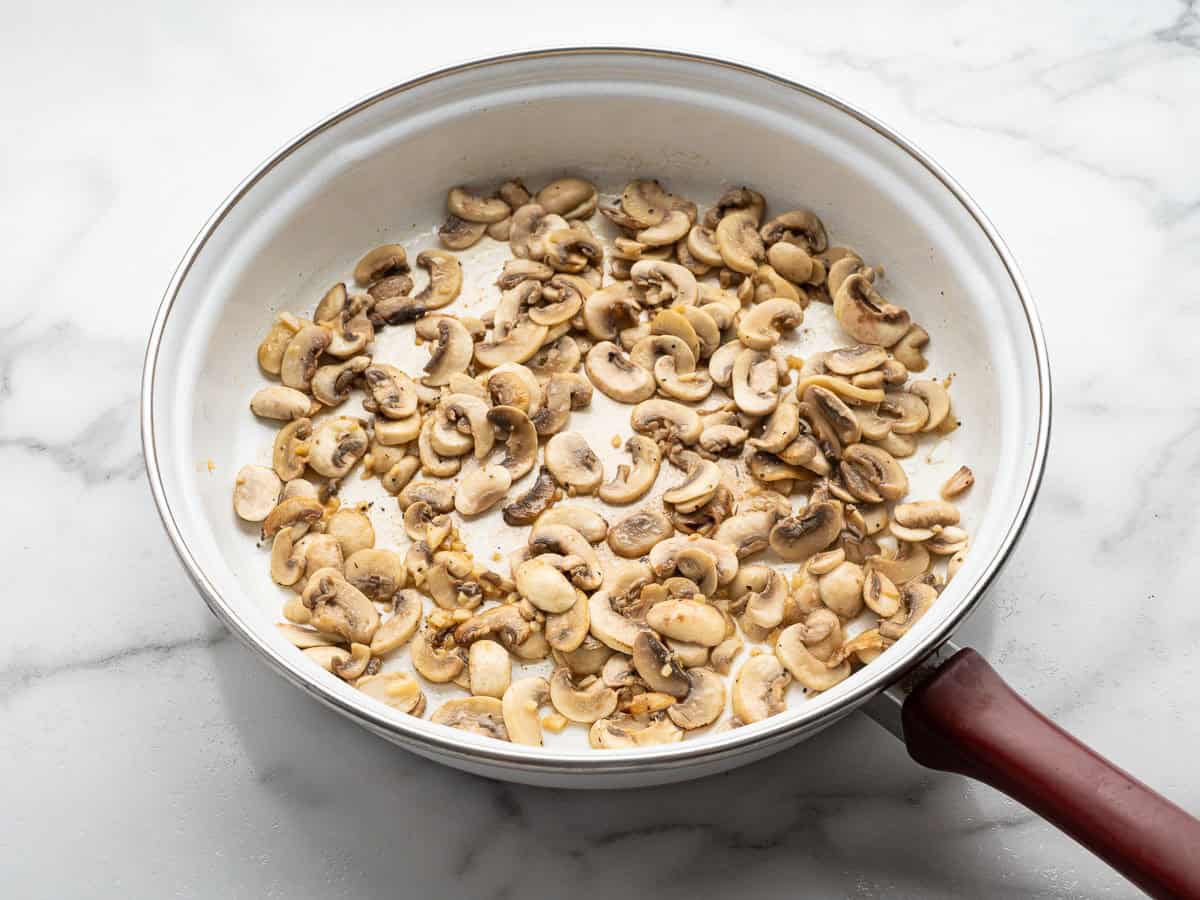 Sliced mushrooms being sautéed in a white frying pan with a brown handle, placed on a white marble countertop—the perfect start for recreating the savory topping found on Alice Springs Chicken.