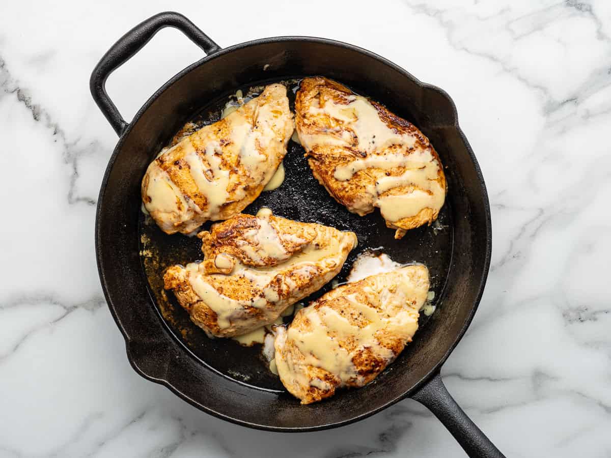 Four grilled chicken breasts topped with melted cheese sit in a black cast iron skillet on a white marble surface, evoking the mouthwatering appeal of classic Alice Springs Chicken.