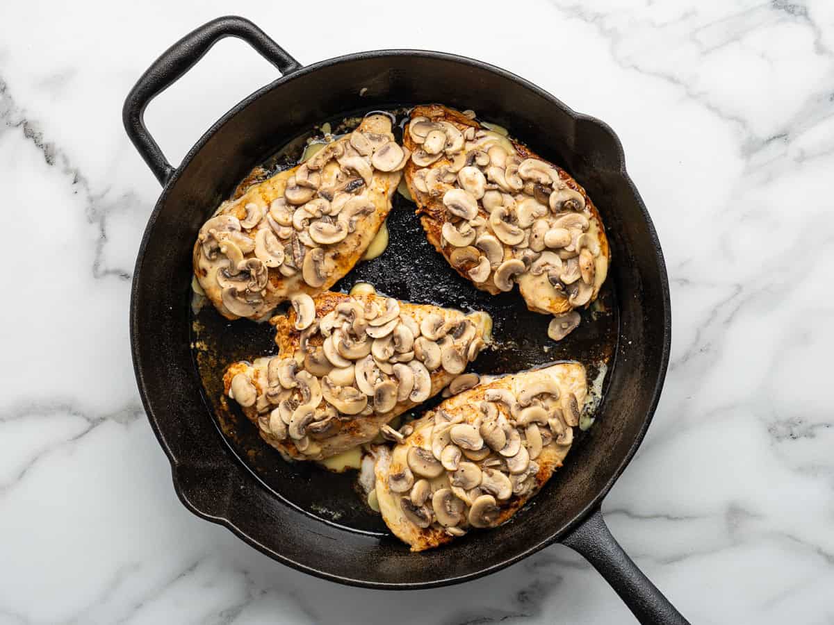 Four pieces of Alice Springs Chicken breast topped with sautéed mushrooms and melted cheese cook in a black cast iron skillet on a white marble surface.