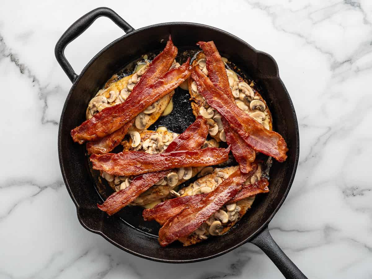 A cast iron skillet with cooked chicken breasts, inspired by the famous Alice Springs Chicken, topped with sautéed mushrooms and crispy bacon strips on a marble surface.