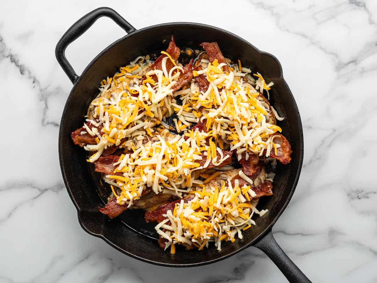 A black cast iron skillet filled with cooked bacon strips and shredded cheese, reminiscent of the classic Alice Springs Chicken toppings, sits on a white marble surface.