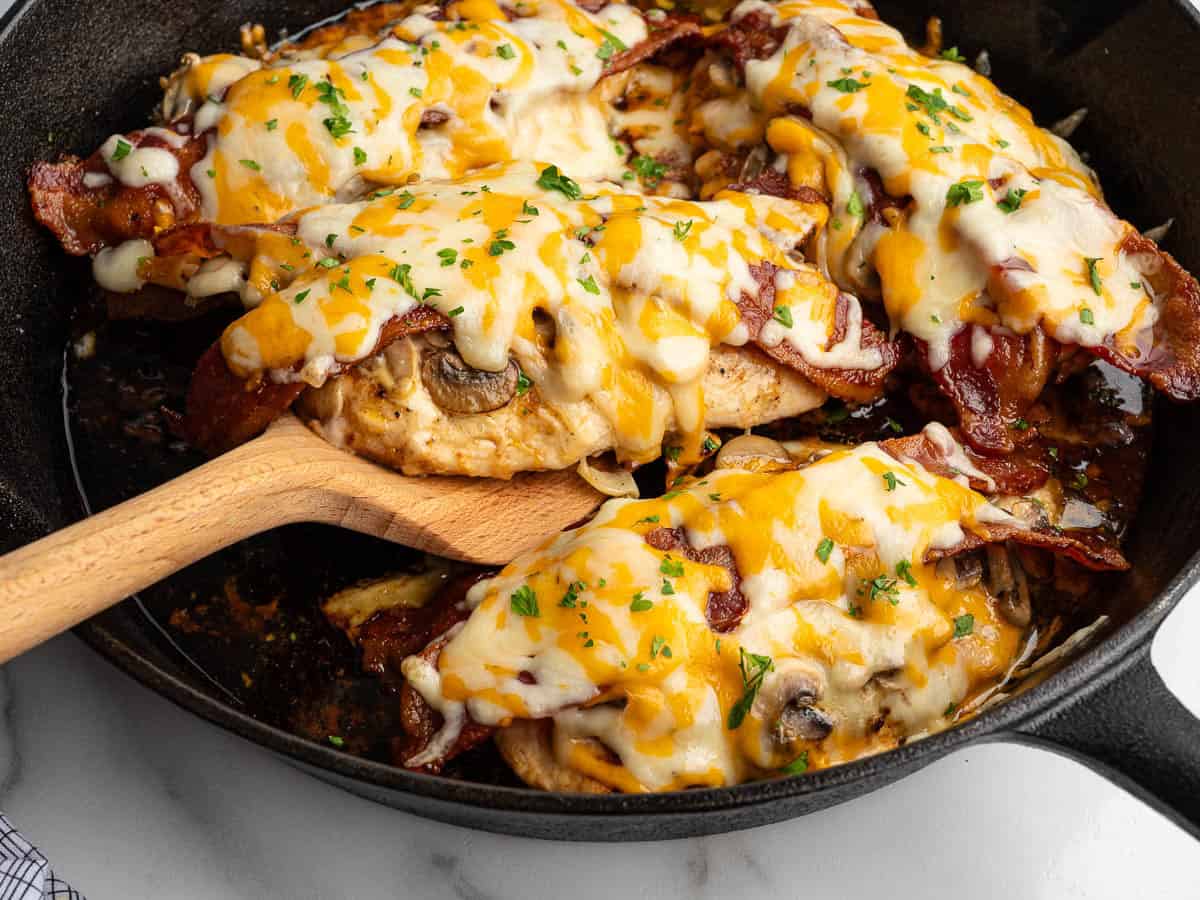 Melted cheese-topped chicken breasts with bacon and mushrooms are in a black skillet, reminiscent of classic Alice Springs Chicken. A wooden spatula lifts one piece; parsley is sprinkled on top.