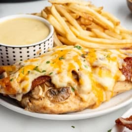 A plate showcasing Alice Springs Chicken—a cheesy chicken breast topped with bacon and mushrooms—served alongside golden French fries and a cup of creamy yellow dipping sauce.
