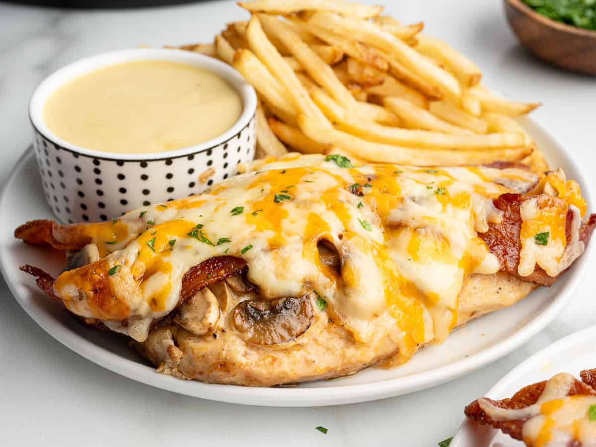 A plate showcasing Alice Springs Chicken—a cheesy chicken breast topped with bacon and mushrooms—served alongside golden French fries and a cup of creamy yellow dipping sauce.