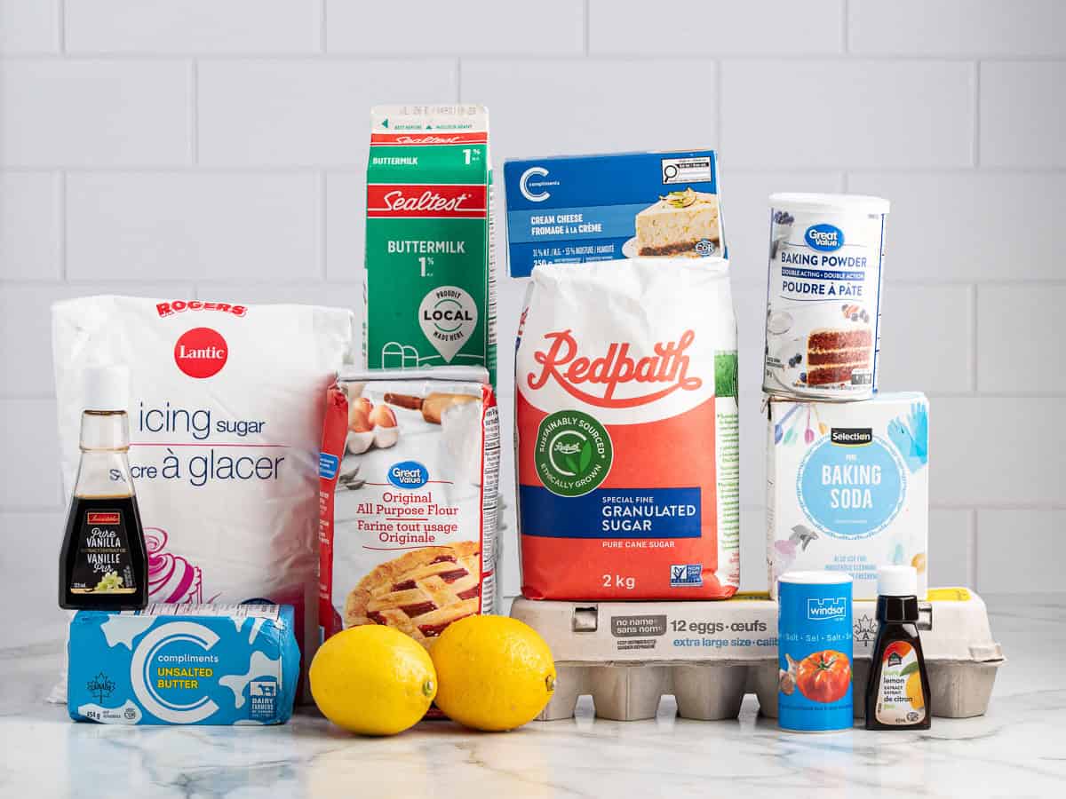Assorted baking ingredients on a counter, including flour, sugar, buttermilk, butter, eggs, lemons, baking powder, baking soda, vanilla extract, and powdered sugar—perfect for creating Lemon Velvet Cake—set against a clean white tiled background.