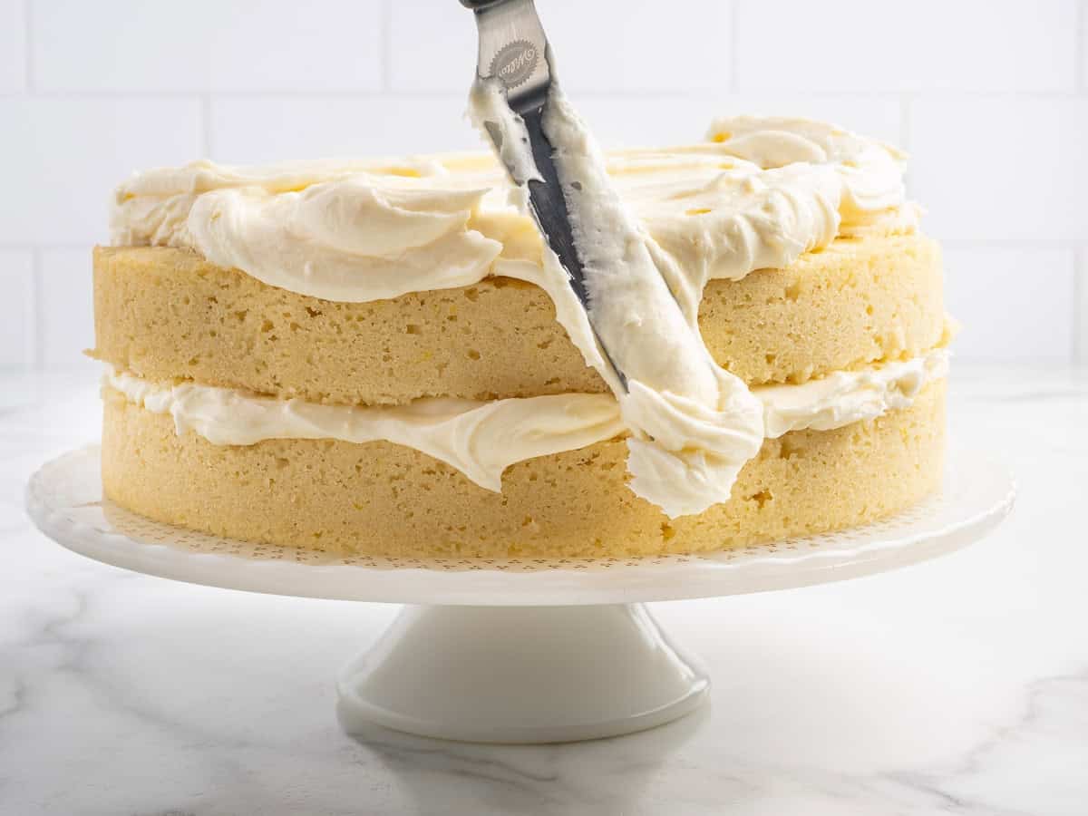 A Lemon Velvet Cake on a white stand is being frosted with a spatula. The cake has two layers with frosting in the middle and on top. The scene is set on a white surface with a white tiled background.