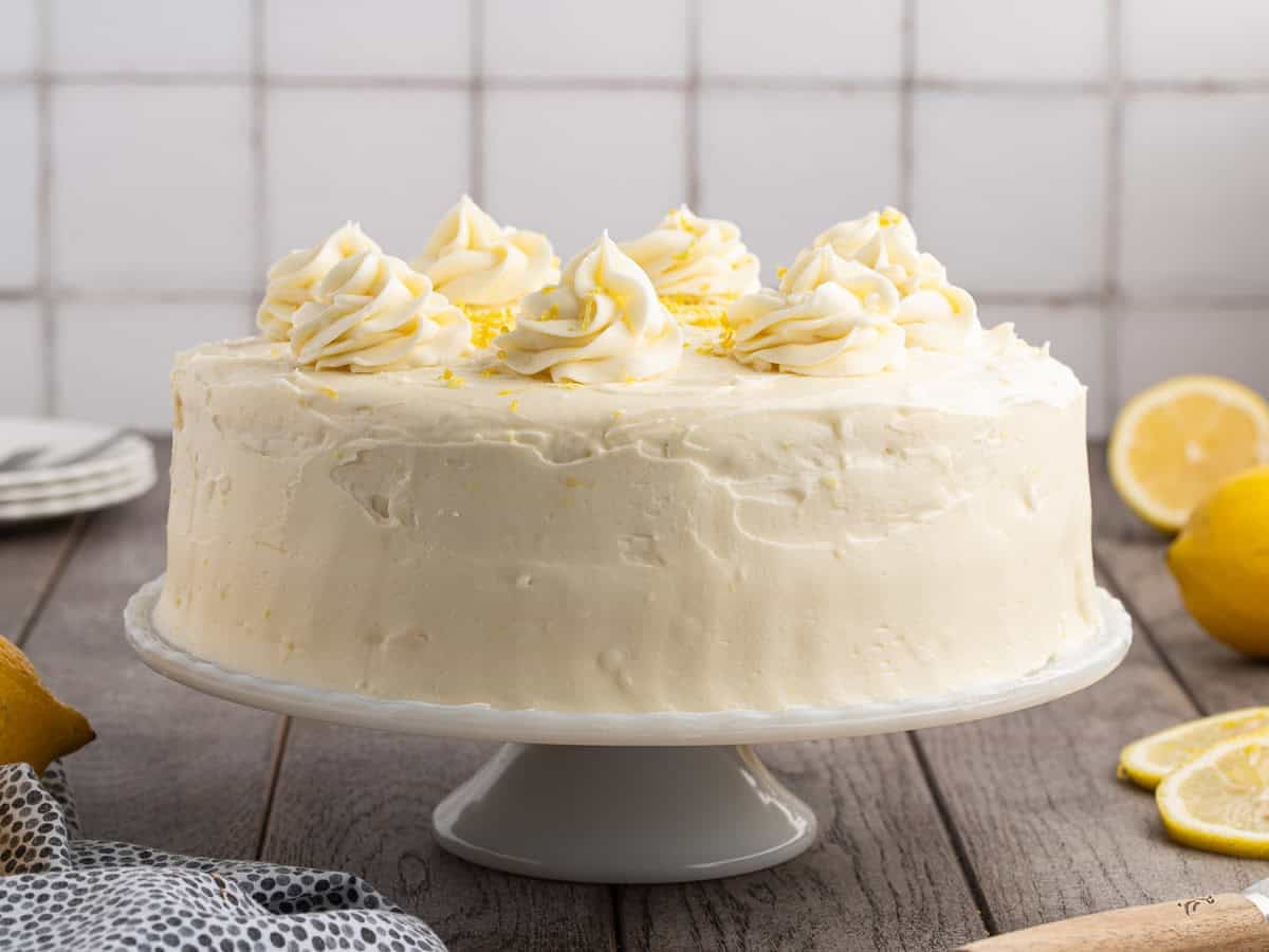A round, frosted Lemon Velvet Cake with swirls of icing on top sits on a white cake stand. Fresh lemons and plates are nearby on a wooden table.