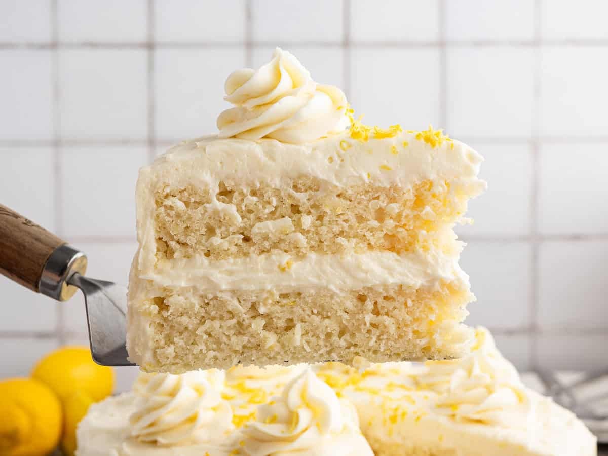 A close-up of a spatula lifting a slice of Lemon Velvet Cake, showcasing its two tender layers with creamy white frosting and bright lemon zest, set against a tiled background.