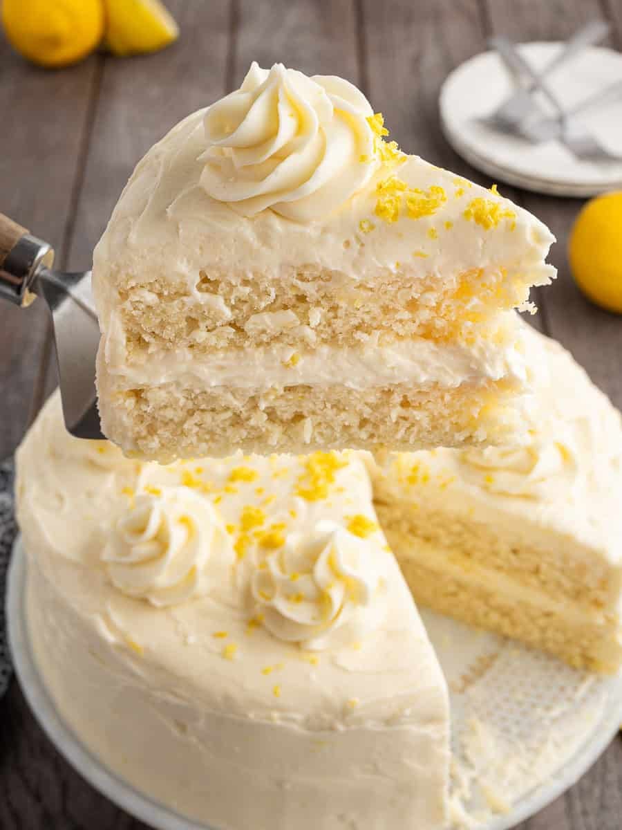 A slice of Lemon Velvet Cake with white frosting and yellow zest is being lifted from a whole cake, which is garnished with piped frosting and lemon zest, on a wooden table.