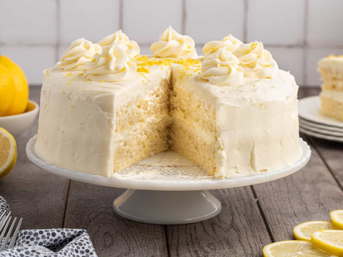 A frosted Lemon Velvet Cake with swirls of icing on top sits on a white cake stand. A large slice is missing. Lemon slices and whole lemons are nearby on a wooden table.
