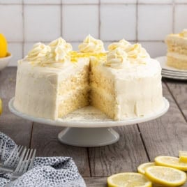 A round Lemon Velvet Cake with white frosting and piped swirls on top sits on a cake stand. A large slice has been removed, revealing the moist, tender layers inside. Lemon slices and a gray napkin are nearby.