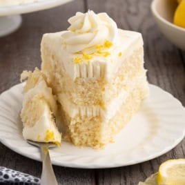 A slice of Lemon Velvet Cake with white frosting sits on a white plate, topped with bright lemon zest. A fork holds a bite of the soft, tender cake. Lemon slices and whole lemons are visible in the background.