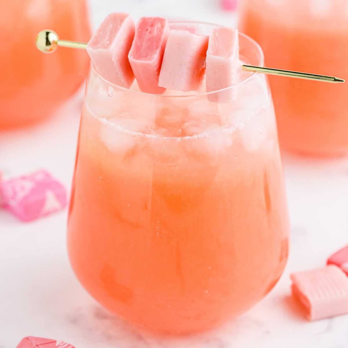Pink cocktail with candy skewer in a stemless glass, surrounded by pink starburst candies on a white surface.