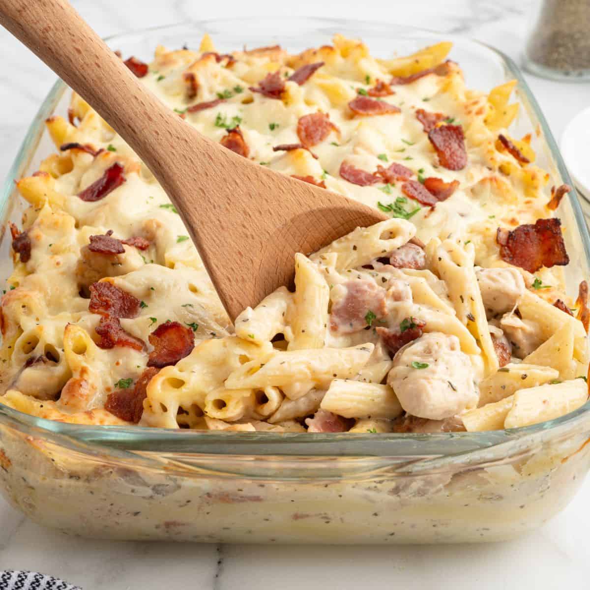 A glass baking dish filled with creamy baked pasta, pieces of chicken, bacon, and cheese. A wooden spoon scoops out a serving of this delicious Chicken Bacon Ranch Casserole. The dish is garnished with chopped herbs.