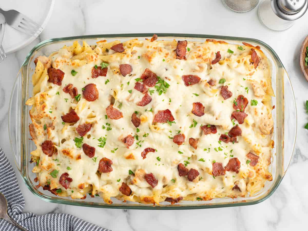 A glass baking dish filled with a hearty Chicken Bacon Ranch Casserole, featuring baked pasta topped with melted cheese, crispy chopped bacon, and sprinkled herbs, placed on a marble surface with a striped towel and dinnerware nearby.