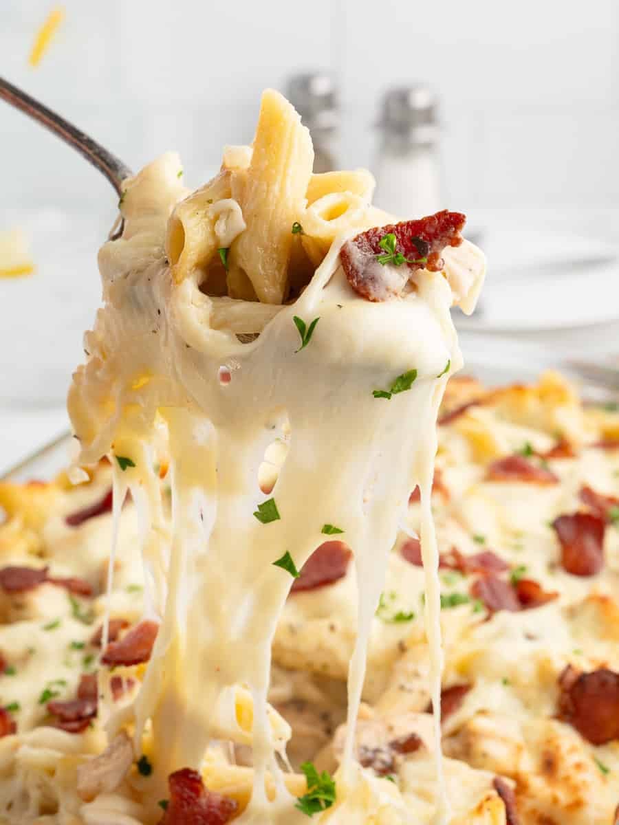 A fork lifts a serving of cheesy Chicken Bacon Ranch Casserole, with melted cheese stretching, bits of bacon, and parsley garnish on top. The background shows more pasta and blurred salt and pepper shakers.