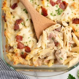 A glass baking dish filled with baked penne pasta, creamy sauce, melted cheese, and bacon makes up this irresistible Chicken Bacon Ranch Casserole. A wooden spoon scoops out a hearty serving, revealing layers of flavor beneath a sprinkle of fresh parsley. Nearby, a striped napkin and sprigs of herbs add the finishing touch to this comforting meal.