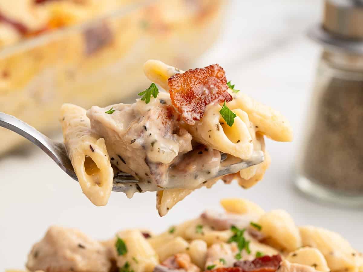 A close-up of a forkful of creamy penne pasta with pieces of bacon, chicken, and parsley highlights the delicious flavors of a Chicken Bacon Ranch Casserole. In the background, a blurred dish of pasta and pepper shaker add to the inviting scene.
